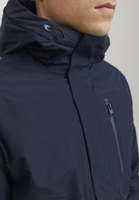 Giacca impermeabile blu navy con colletto alto, cordino regolabile e tasca sul petto con zip. Texture liscia, design elegante.