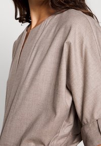 Blouse en tissu doux couleur brun clair, dotée d'un décolleté en V, de manches longues avec poignets et de détails de couture subtils le long des coutures.