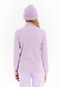 Asmuo vilkintis šviesiai purpurinį raštuotą ilgų rankovių marškinį, atitinkančią megztą kepurę ir šviesiai purpurines kelnes, atsisukęs nugarą prieš baltą foną.