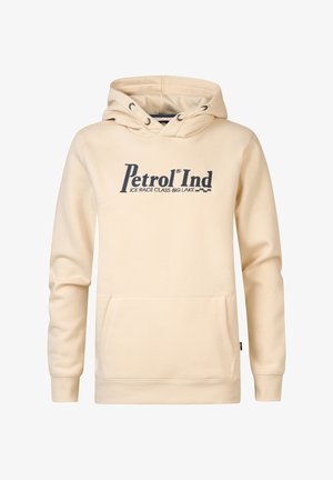 Dziecko ubrane w beżową bluzę z kapturem marki Petrol Ind, niebieskie dżinsy oraz czarno-białe trampki, stojące na jednolitym tle.