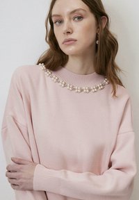 Pulover roz deschis cu o guler texturat, având perle decorative și strasuri pe colier. Croială supradimensionată cu mâneci lungi.