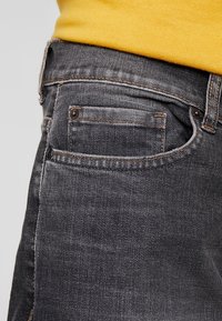 Lähikuva mustista denim-farkuista, joissa on kupariset niitit, kontrastina sinapinkeltainen neulepaita, joka on työnnetty sisään.