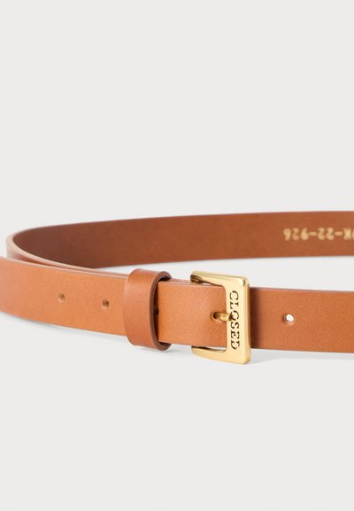 Ceinture en cuir fauve avec boucle rectangulaire dorée gravée de "CLOSED" et plusieurs trous de réglage sur fond blanc.