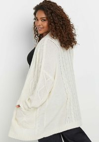 Langer, grob gestrickter Cardigan in Off-White, mit offenem Frontbereich, strukturierten Mustern und gerippten Bündchen. Weicher Stoff, entspannter Schnitt und oversized Design.