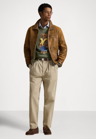 Καφέ δερμάτινο μπουφάν πάνω από πράσινο πουλόβερ με σχέδιο αρκούδας, συνδυασμένο με μπεζ παντελόνια και καφέ παπούτσια με πορτοκαλί κορδόνια.