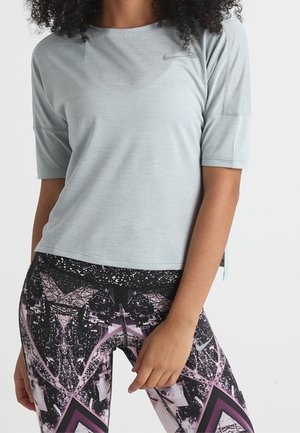 Personne portant un haut à manches courtes gris clair et un legging à motif géométrique noir, blanc et rose sur fond blanc uni.