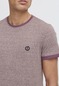Camiseta de manga corta en tejido marrón jaspeado con un cuello y puños de canalé morados, que presenta un pequeño logo azul marino en el pecho.