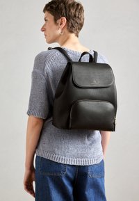 Zaino in pelle nera con una forma strutturata, patta superiore, tasca frontale con cerniera e spallacci regolabili. Superficie testurizzata con un marchio discreto.