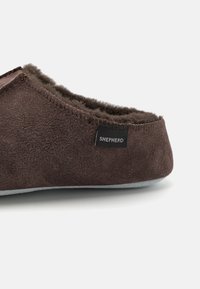 Chausson en daim marron avec une doublure intérieure douce, arborant une étiquette noire portant la mention "SHEPHERD" et une semelle grise.