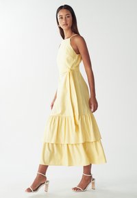 Gele linnen jurk met een aansluitend lijfje, verstelbare bandjes en een gelaagde, gerimpelde zoom. Draagt met witte hoge hakken.