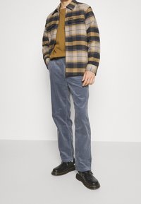 Kostkovaná flanelová košile v béžové, námořnické a žluté barvě; hořčicový svetr; modré kordové kalhoty; černé boty se žlutým prošíváním.