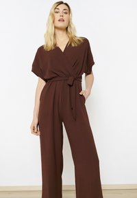 Bruine wikkeljumpsuit met korte mouwen, een strik taille en wijde pijpen. Gemaakt van lichtgewicht stof met een gladde textuur.