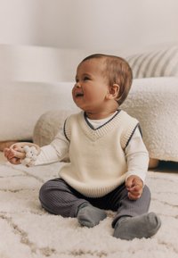 Infant wearing a cream knitted vest with navy accents, gray ribbed pants, and gray socks, holding a wooden teething ring with beads.