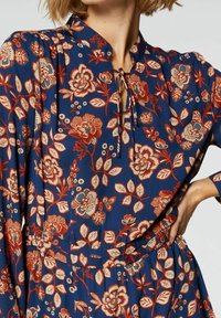 Kwiecista sukienka w granatowym kolorze, z czerwonymi i beżowymi wzorami. Posiada kołnierz mandarynkowy, detal w postaci kokardy przy dekolcie oraz długie rękawy.