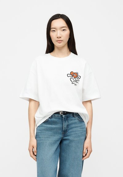 Femme aux longs cheveux foncés portant un t-shirt blanc oversize avec un imprimé graphique coloré et un jean bleu, debout sur fond blanc.