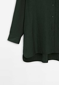 Tummanvihreä, pitkähihainen paita, joka on valmistettu teksturoidusta kankaasta. Siinä on napillinen etuosa ja kaareva helma, jossa on hieman läpikuultava alaosa.