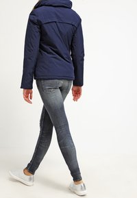 Giacca a soffietto blu navy con colletto alto, jeans slim fit in lavaggio medio e scarpe slip-on argentate. Vista da dietro, che mostra l'outfit in movimento.