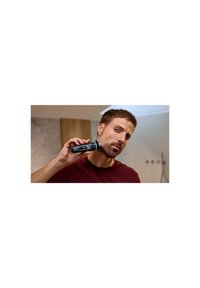 Elektrisch scheerapparaat met een zwart en zilver ontwerp, met een ronde kop. De gebruiker houdt het handvat vast terwijl hij gezichtsbeharing scheert; de achtergrond is neutraal.