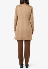 Cappotto in misto lana color tan con design sartoriale, maniche lunghe e dettaglio a bottone sui polsini. Abbinato a pantaloni attillati marroni e scarpe nere.
