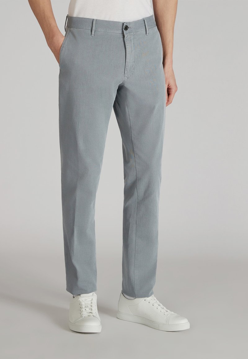 Pantaloni grigi chiari realizzati in un tessuto liscio, con un taglio classico, passanti per la cintura e chiusura con un singolo bottone. Abbinati a sneakers bianche.