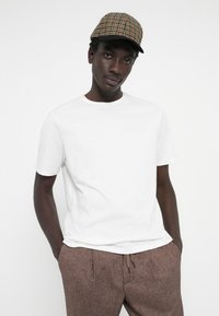 White cotton t-shirt with short sleeves, paired with brown patterned pants. The model wears a green and brown checkered cap.