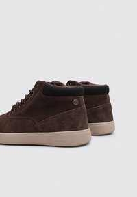 Zapatos de ante marrón de caña alta con un acabado texturizado, puntera redondeada, cuello acolchado negro y suela de goma beige; se observa un detalle mínimo de marca.