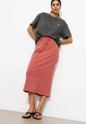 Femme appuyée contre un mur blanc, portant un t-shirt oversize gris foncé, une jupe midi corail avec cordon de serrage et des sandales noires à bout ouvert.