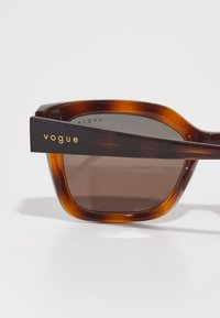 Ochelari de soare tortoiseshell maro cu formă unghiulară și lentile degradé. Bratul are un logo auriu "vogue". Textură din plastic neted.