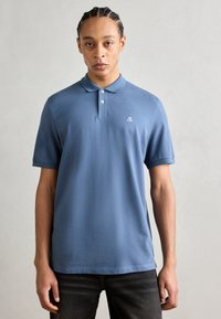 Polo bleu din bumbac, cu guler clasic, mâneci scurte și un mic logo pe piept. Textură simplă și netedă.