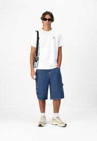 T-shirt blanc à manches courtes avec logo, shorts en denim bleu, et baskets beiges avec des chaussettes blanches. Des lunettes de soleil et un sac bandoulière complètent le look.
