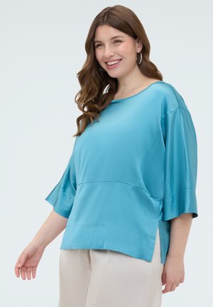 Jeune femme aux longs cheveux bruns portant des boucles d'oreilles argentées en forme d'anneau, un chemisier ample en satin turquoise et un pantalon beige, souriante et posant de profil.