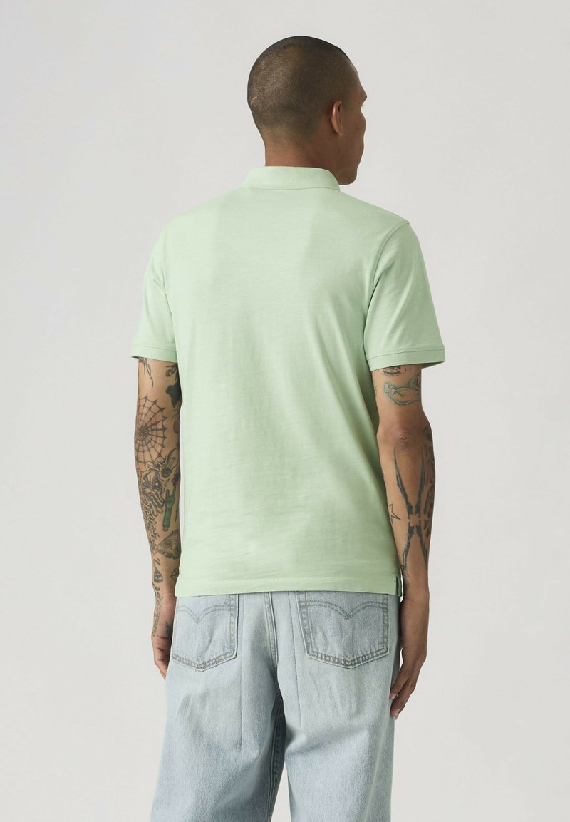 Man met getatoeëerde armen, gekleed in een lichtgroen poloshirt met korte mouwen en lichtblauwe spijkerbroek, gezien van achteren tegen een effen achtergrond.