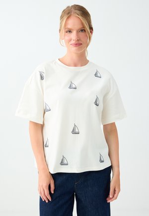 Femme aux cheveux blonds portant une chemise blanche à manches courtes avec de petits motifs noirs de voiliers et un jean bleu foncé sur un fond uni.