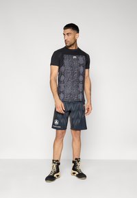 Μαύρο πουκάμισο με κοντά μανίκια και σχέδιο φιδιού, συνδυασμένο με σκούρα σορτς με σχέδιο κύματος. Μαύρα και ταμπά αθλητικά παπούτσια ολοκληρώνουν την εμφάνιση.