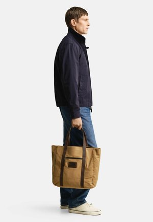 Man draagt een donker jack en blauwe jeans, houdt een beige canvas tas met donkere handgrepen vast en staat tegen een witte achtergrond.