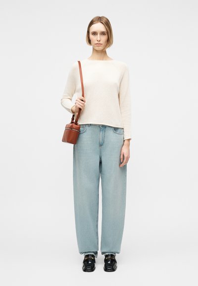 Γυναίκα που φοράει λευκή μπλούζα με μακριά μανίκια, ανοιχτό μπλε φαρδιά τζιν, μαύρα παπούτσια, κρατώντας μια μικρή καφέ δερμάτινη τσάντα ώμου σε λευκό φόντο.