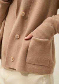 Cárdigan marrón de punto con textura acanalada, que presenta tres botones y un bolsillo frontal. Tejido suave con un corte relajado, sin patrones visibles.