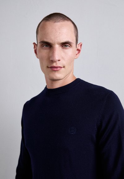 Man with short hair wearing a dark blue crewneck sweater, facing forward against a plain light gray background.
