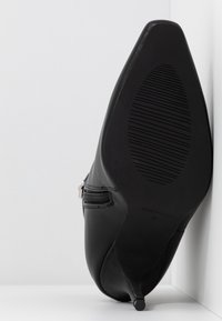 Schwarze, lederne, spitz zulaufende Ankle-Boots mit schlankem Absatz, Reißverschluss an der Seite und strukturiertem Sohlen, vor einer weißen Eckwand platziert.