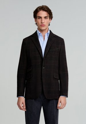 Blazer marrón a cuadros hecho de tela texturizada, con solapas de muesca, dos bolsillos frontales y cierre de un solo botón. Llevado sobre una camisa azul clara.