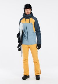 Heren snowboardoutfit met een blauwe en gele jas en oranje broek; materialen lijken waterdicht, met zwarte handschoenen en stevige zwarte laarzen.