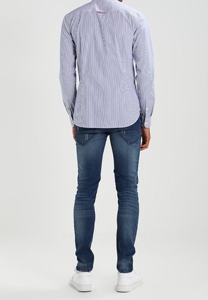 Mies, jolla on vaaleansininen raidallinen pitkähihainen paita, tummansiniset slim fit -farkut ja valkoiset lenkkarit, seisoo selin yksivärisellä taustalla.
