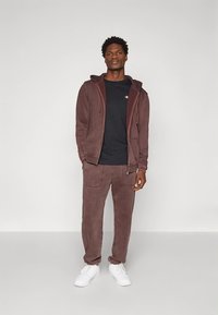 Felpa con cappuccio color marrone scuro e pantaloni jogger coordinati in tessuto morbido, caratterizzati da una tasca con cerniera e cuciture a contrasto. Scarpe da ginnastica bianche completano il look.