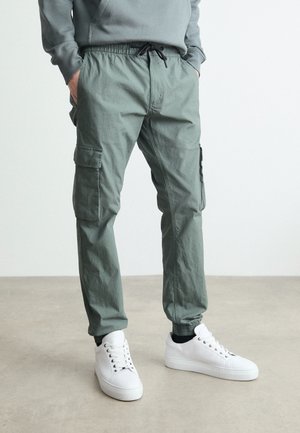 Grüne Cargo-Hosen mit einem elastischen Bund und Kordelzug, ausgestattet mit Seitentaschen und schmal zulaufenden Beinen, kombiniert mit weißen Sneakers.