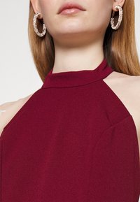 Burgundy halter neck top with a smooth texture, featuring cut-out shoulder details and a clean, minimalistic design. Large hoop earrings visible.