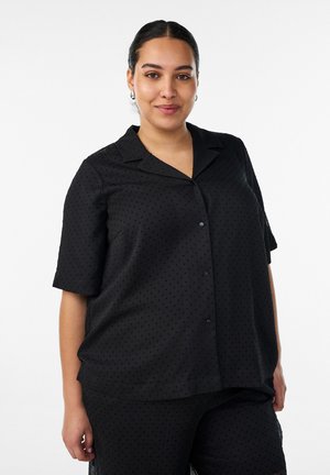 Frau mit dunklem Haar in einem niedrigen Dutt trägt ein schwarzes kurzärmeliges Hemd mit dezenten Punkten und passende schwarze Hose, lächelt leicht.
