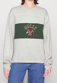 Šedá mikina má zelený horizontální panel s vyšívaným textem "GUESS" a designem jelena; široké rukávy a volný střih.