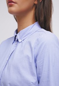 Chemise bleu clair à boutons avec col structuré et manches retroussées, présentant une texture lisse et des détails de couture subtils.