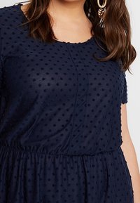 Marinblå klänning med en strukturerad tyg med upphöjda prickar, korta ärmar och ett elastiskt midjeband för en avslappnad passform.