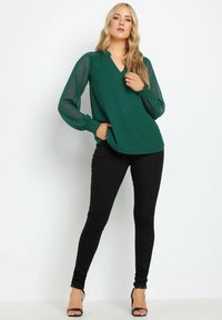 Blusa verde a scollo a V con tessuto testurizzato, maniche trasparenti e polsini arricciati. Abbinata a jeans skinny neri e sandali con tacco neri.
